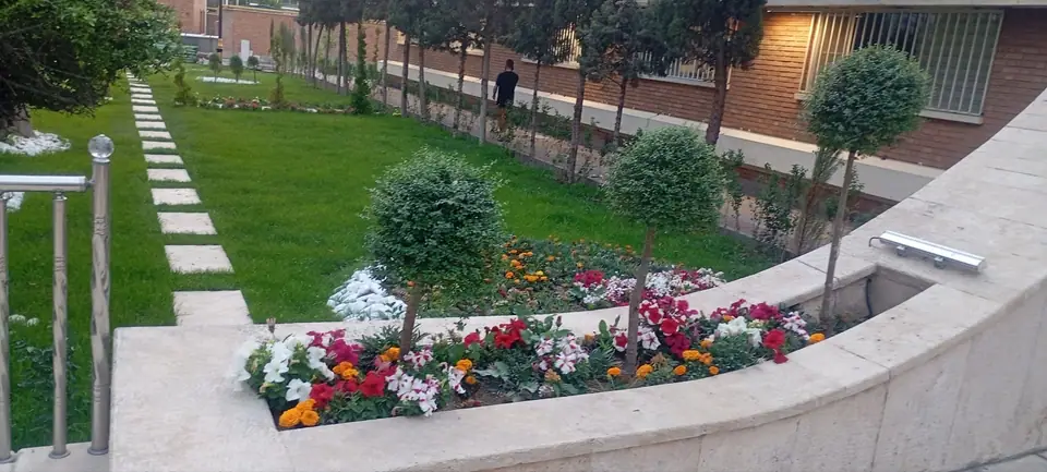 درختچه‌های فرم‌داده‌شده و باغچهٔ گل رنگارنگ کنار دیوارهٔ سنگی